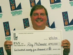 Man with mustache smiles with oversized check in front of a white background patterned with the 幸运168®飞艇开奖预测网-Louisiana全国统一历史记录预测_在线查询开奖最新数据 logo