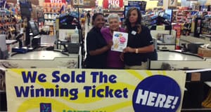 Three women hold an Easy 5 winner sign while standing behind a retail counter with a banner that reads "We Sold the Winning Ticket Here!"