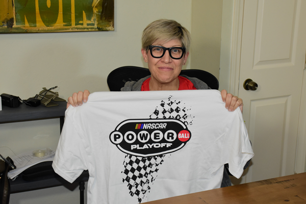Woman with short blonde hair and glasses holds a white tshirt emblazoned with the Nascar Powerball Powerplay logo and tire treads