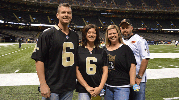 Two men and two women in Saints gear smile together on the sidelines of a game