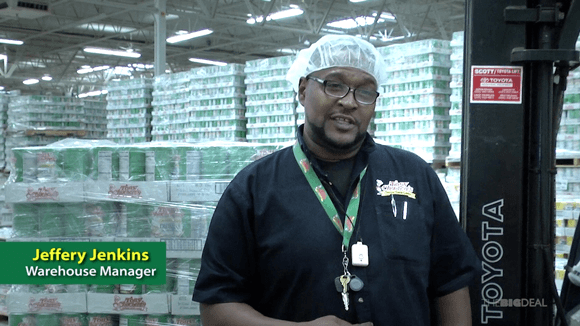 Man in a black polo, lanyard, and hair net talks to the camera in a warehouse.