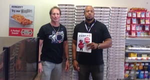 Two men pose in a retailer, one holding a Powerball winner sign.