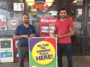 Two men stand outside of a retailer and hold a sign that reads "We Sold the Winning Ticket Here!"
