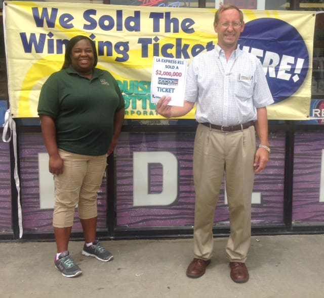Man holds a winner sign and smiles with a woman in front of a storefront while standing in front of a yellow banner that reads "We Sold the Winning Ticket Here!"