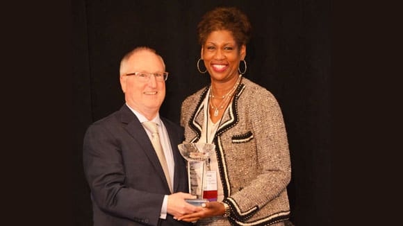 Man and woman in business attire pose with a glass award in front of a black background