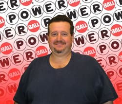 Man in a black tshirt smiles in front of a red and white background branded with the Powerball logo