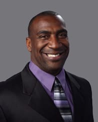 Headshot of a man with a crew cut in a black suit jacket, purple shirt, and black and white striped tie smiling in front of a gray background