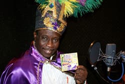 Man in a purple silk cape and towering mardi gras feathered hat holds a scratch off ticket and smiles seductively at the camera