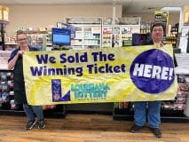 Two people stand in front of a counter and hold a large yellow banner that reads "We Sold the Winning Ticket Here"