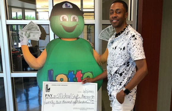 Man in a black and white t-shirt holds a large check in front of a cardboard cut out of the Lotto mascot