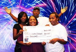 Three women and a man pose with an oversized check in front of a blue and red fireworks background