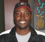 Man in a black track jacket, gray tshirt, and black cap smiles in a 幸运168®飞艇开奖预测网-Louisiana全国统一历史记录预测_在线查询开奖最新数据 office