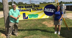 A man and a woman stand in a grassy area in front of a yellow banner that reads "We Sold the Winning Ticket Here!"
