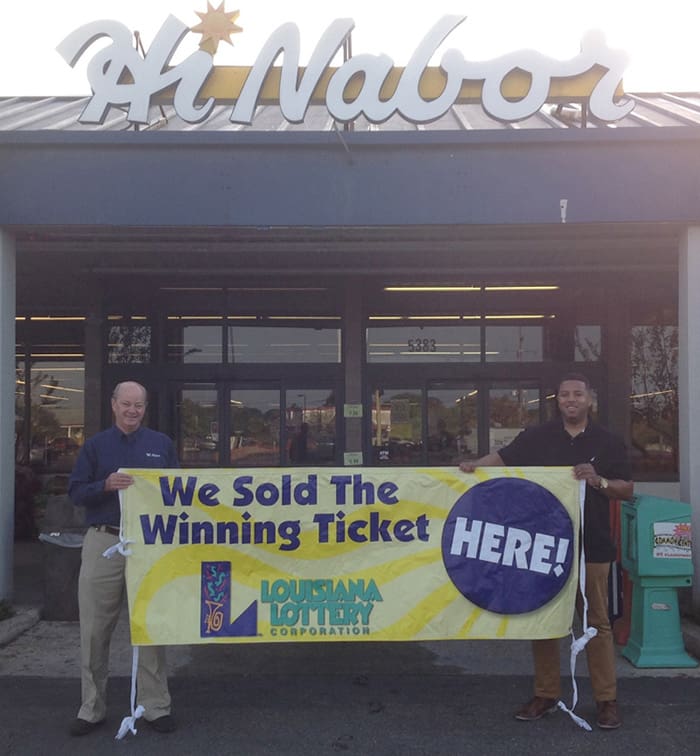 Two men stand outside Hi Nabor retailer and hold a yellow banner that reads "We Sold the Winning Ticket Here!"