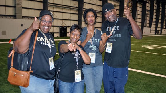 Saints fans smile, give thumbs up, and point at the camera on an indoor turf field