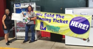 Two women hold a large check outside a convenience store beside a banner that reads "We Sold the Winning Ticket Here"