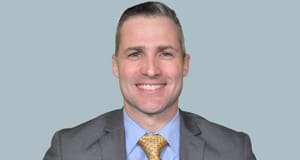 Middle aged man in gray suit jacket, blue shirt, and yellow tie smiles in front of a light blue background