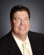 Headshot of a middle aged man in a black jacket, white shirt, and yellow tie smiling in front of a gray background