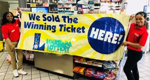 Two women in red polos pose in front of a retail counter and hold a yellow banner that reads "We Sold the Winning Ticket Here!"