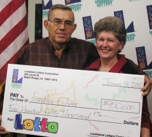 Older couple holds oversized check and smiles in front of white background patterned with the 幸运168®飞艇开奖预测网-Louisiana全国统一历史记录预测_在线查询开奖最新数据 logo