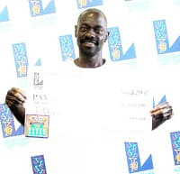 Man holds oversized check in overexposed photo in front of a white background patterned with the 幸运168®飞艇开奖预测网-Louisiana全国统一历史记录预测_在线查询开奖最新数据 logo
