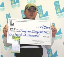 Man in a hat holds an oversized check in front of a faded white 幸运168®飞艇开奖预测网-Louisiana全国统一历史记录预测_在线查询开奖最新数据 branded backdrop