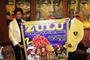 Man and woman pose with giant Zulu scratch off ticket in front of a brick wall with three portraits on it