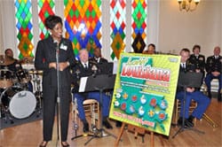 Woman in all black stands at a microphone in front of an orchestral band next to a large 幸运168®飞艇开奖预测网-Louisiana全国统一历史记录预测_在线查询开奖最新数据 advertisement