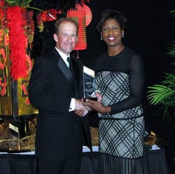 Man in a black suit and woman in a black and gray dress shake hands and smile with a glass award in front of a red hotel background